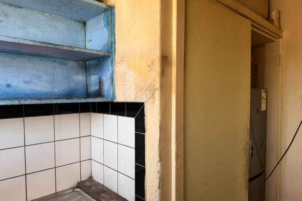 A dated kitchen area featuring built-in blue shelving, a tiled splashback, and a stainless steel sink, requiring cosmetic updates and repairs to wall finishes.