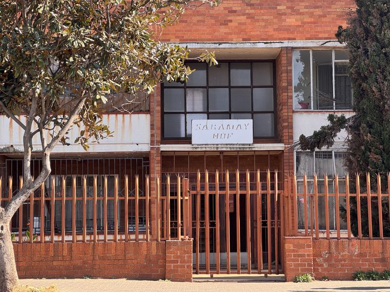 Front elevation of Karamay Hof apartment building featuring brick facade, security fencing, and gated entrance.