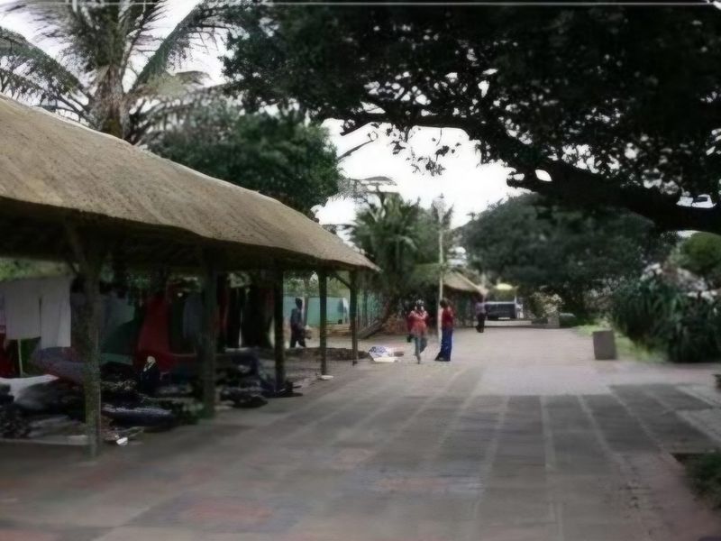 An outdoor area featuring a thatched roof structure and a paved walkway, surrounded by mature trees and landscaping.