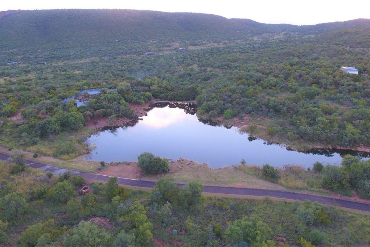 An aerial view of the vacant residential land featuring a natural dam, paved access road, and surrounding indigenous bushveld landscape.