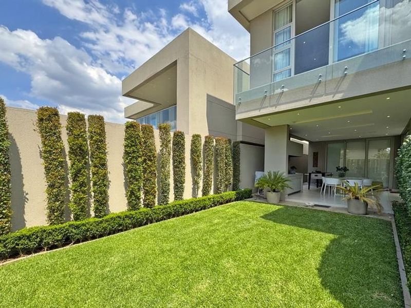 A well-maintained rear garden featuring a manicured lawn, tall privacy hedges, and a covered patio area with a built-in braai and balcony above.