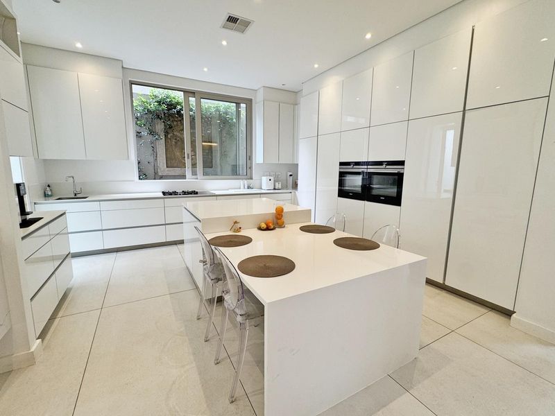 Modern, high-end kitchen featuring sleek white cabinetry, a large central island with integrated seating, double wall ovens, and premium stone countertops.