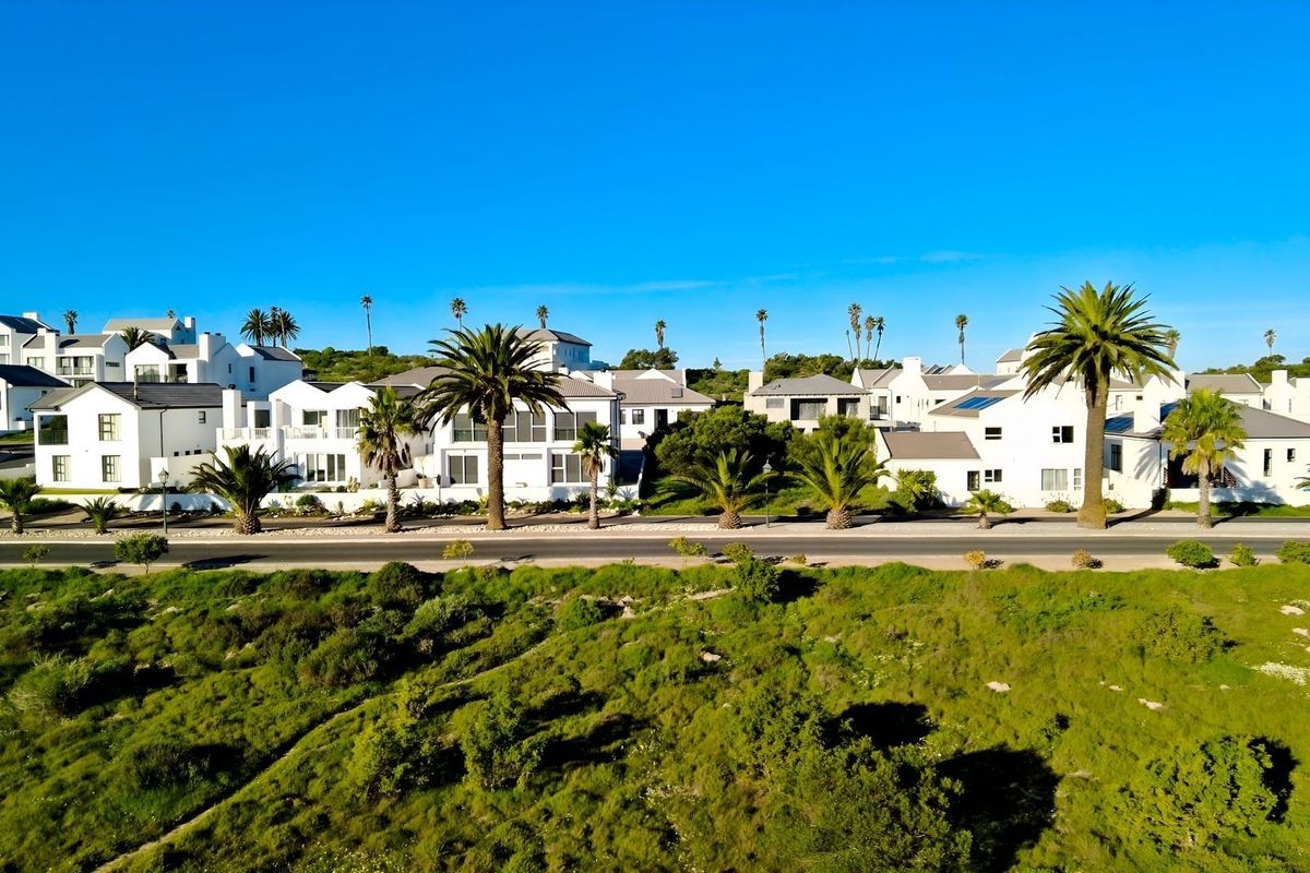 An aerial view of a residential estate featuring white modern homes, palm trees, and a paved road set against a clear blue sky.