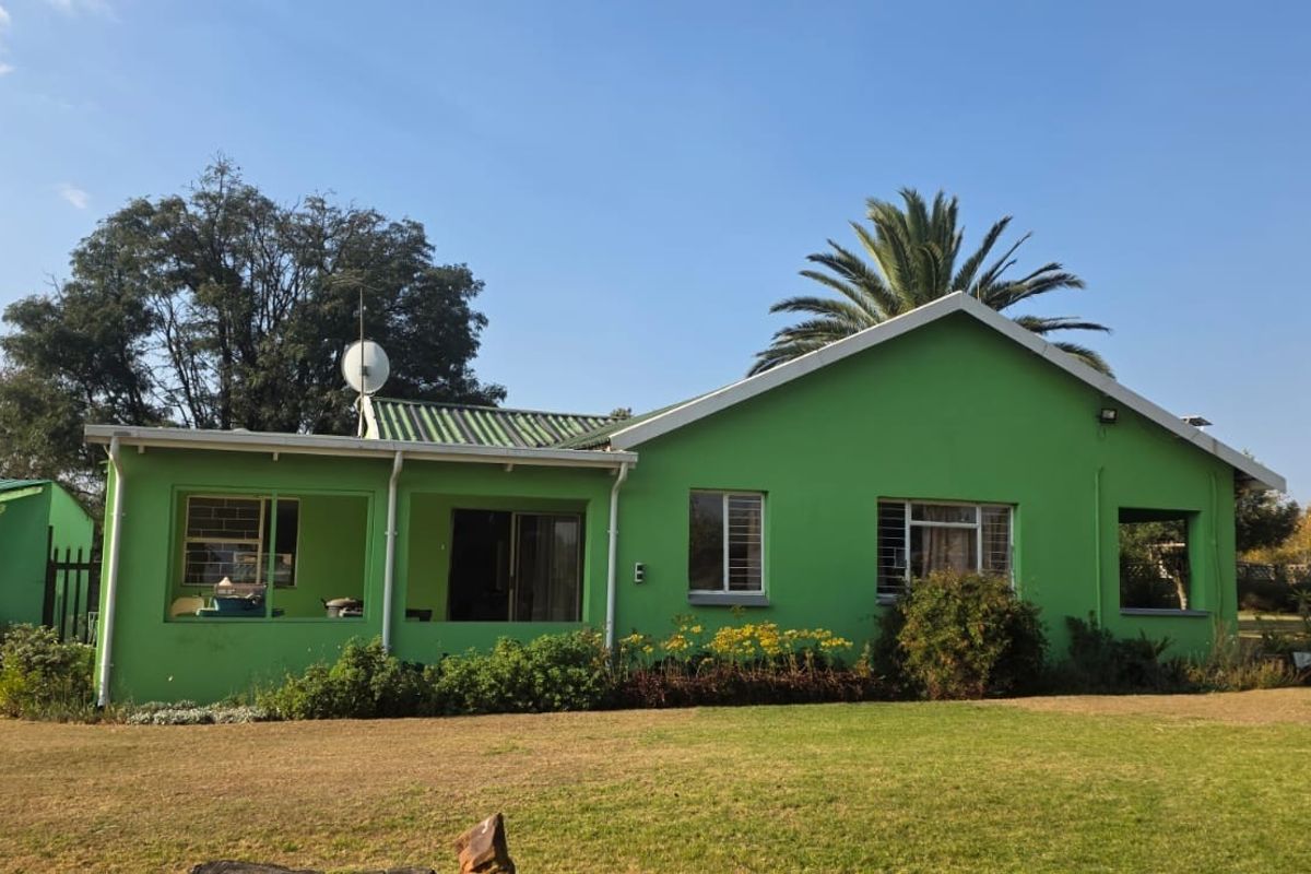 Rear view of a farm-style house featuring a green exterior, corrugated metal roof, and a covered patio area set within a large garden.