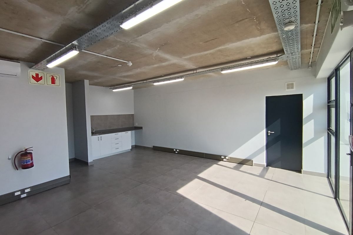 Modern open-plan industrial office space featuring exposed concrete ceilings, tiled flooring, a compact kitchenette, and integrated lighting systems.