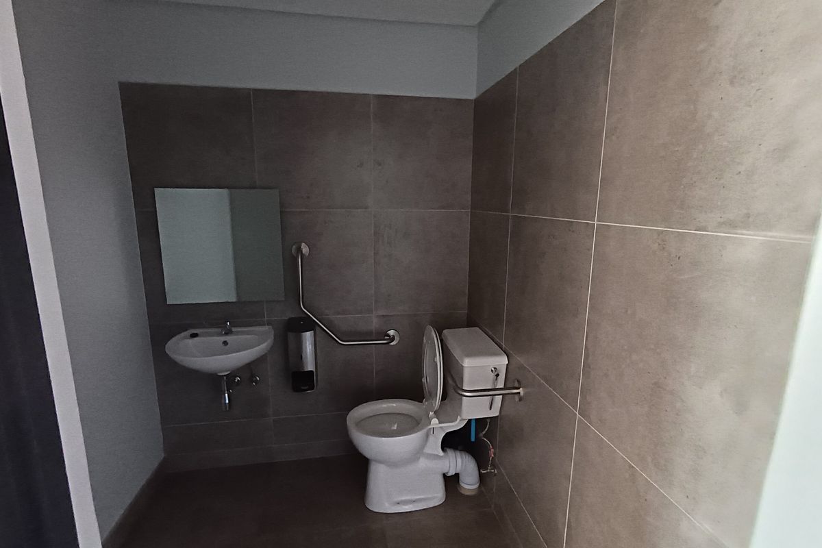 A functional industrial-style accessible bathroom featuring tiled walls, a wall-mounted basin, toilet, and safety grab rails.