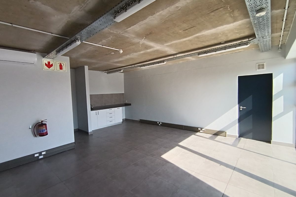Open-plan industrial office space featuring exposed concrete ceilings, tiled flooring, a small kitchenette, and integrated air conditioning.