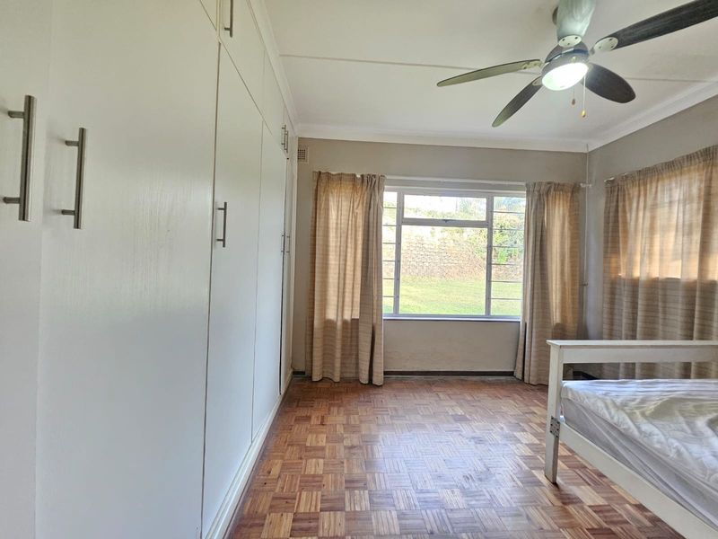 A bedroom featuring classic parquet flooring, extensive built-in cupboards, a ceiling fan, and a large window providing natural light.