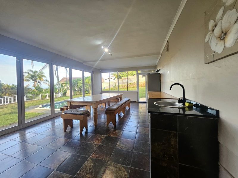 Open-plan living and dining area featuring tiled floors, a kitchenette counter with sink, and large sliding glass doors overlooking the pool and garden.