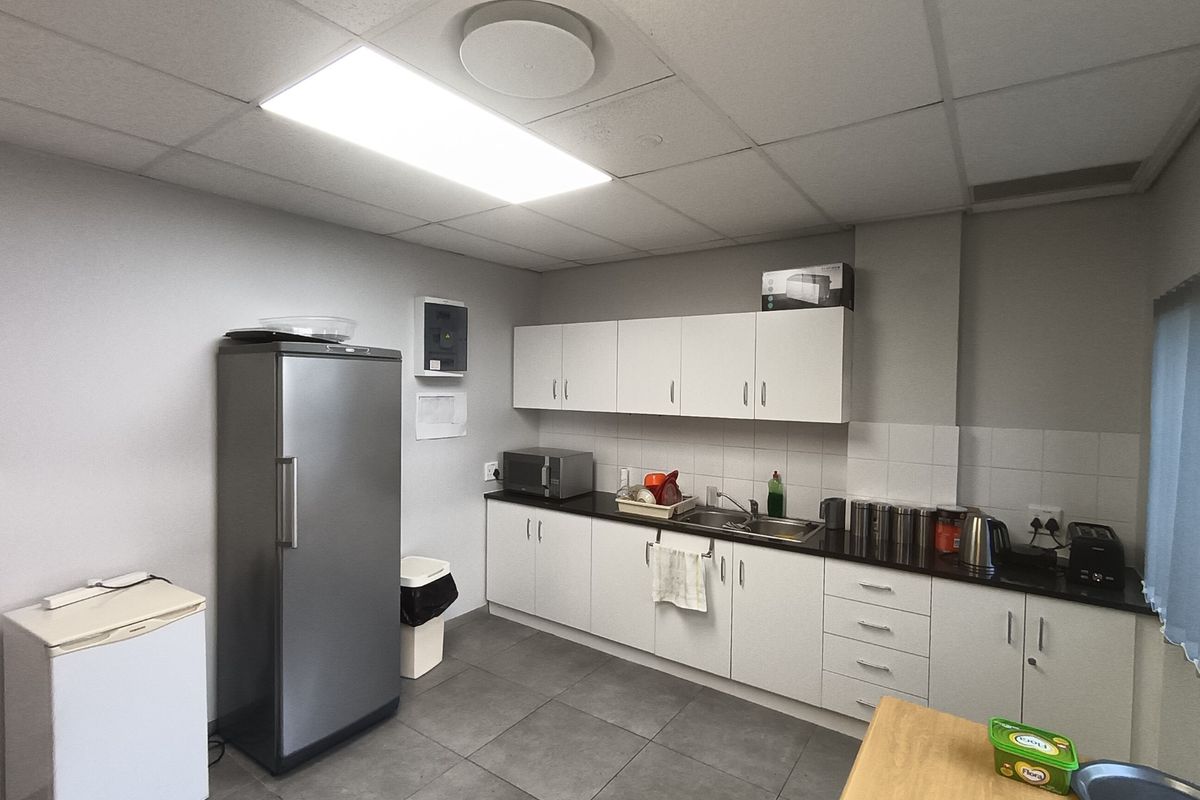 A functional office kitchenette featuring white cabinetry, dark countertops, a stainless steel sink, and tiled flooring under a drop ceiling.