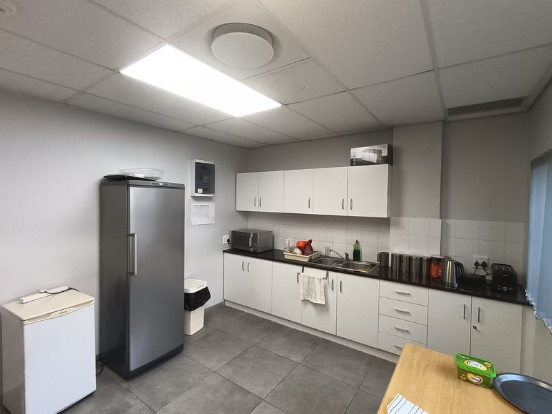 A functional office kitchenette featuring white cabinetry, dark countertops, a stainless steel sink, and tiled flooring under a drop ceiling.