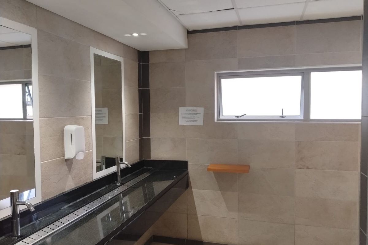 A clean, functional office bathroom featuring a long granite vanity with integrated basins, tiled walls, and a drop ceiling.