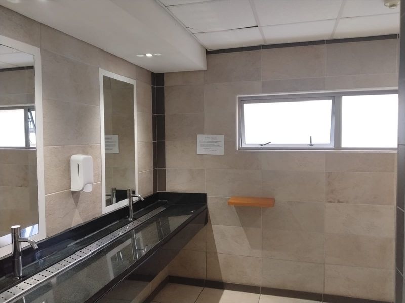 A clean, functional office bathroom featuring a long granite vanity with integrated basins, tiled walls, and a drop ceiling.