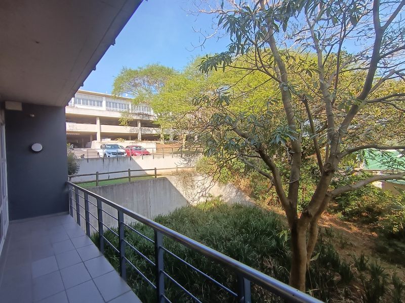 A covered balcony featuring tiled flooring and a metal balustrade, overlooking a landscaped garden area and adjacent parking structure.
