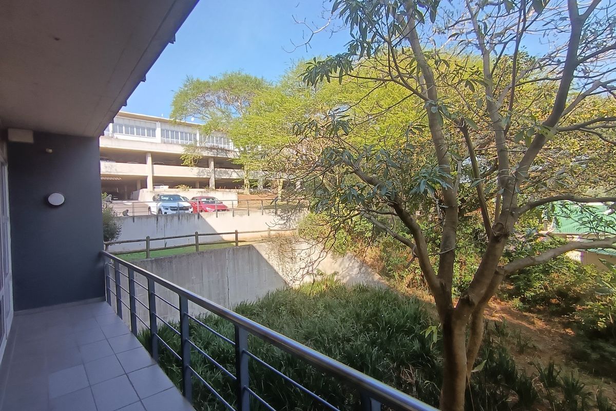 A covered balcony featuring tiled flooring and a metal balustrade, overlooking a landscaped garden area and adjacent parking structure.