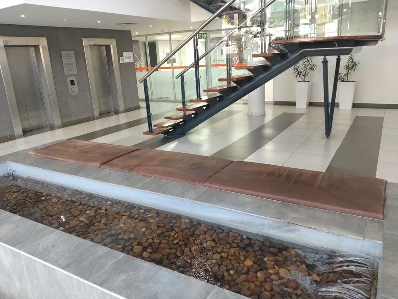 Modern office building lobby featuring a floating staircase with glass balustrades, dual elevators, and a decorative indoor water feature with stone detailing.