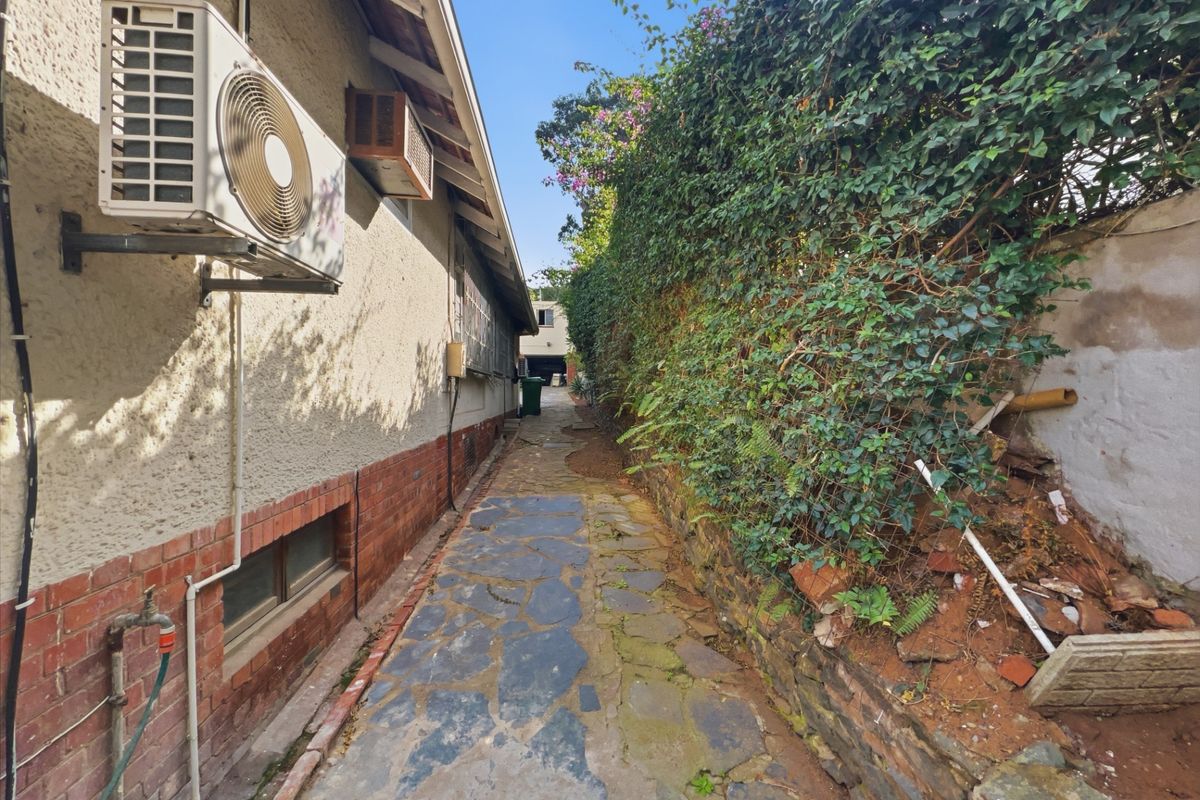 A narrow side passage featuring a stone paved walkway, exterior air conditioning unit, and a retaining wall with overgrown greenery.