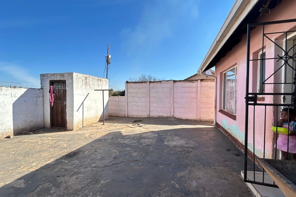A rear exterior area featuring a concrete paved yard, enclosed by boundary walls with security features and visible wear to the building facade.