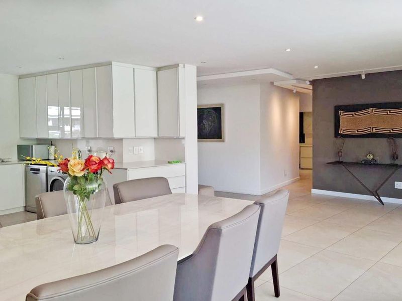 A bright open-plan living and dining area featuring modern white kitchen cabinetry, recessed lighting, and neutral tiled flooring throughout.