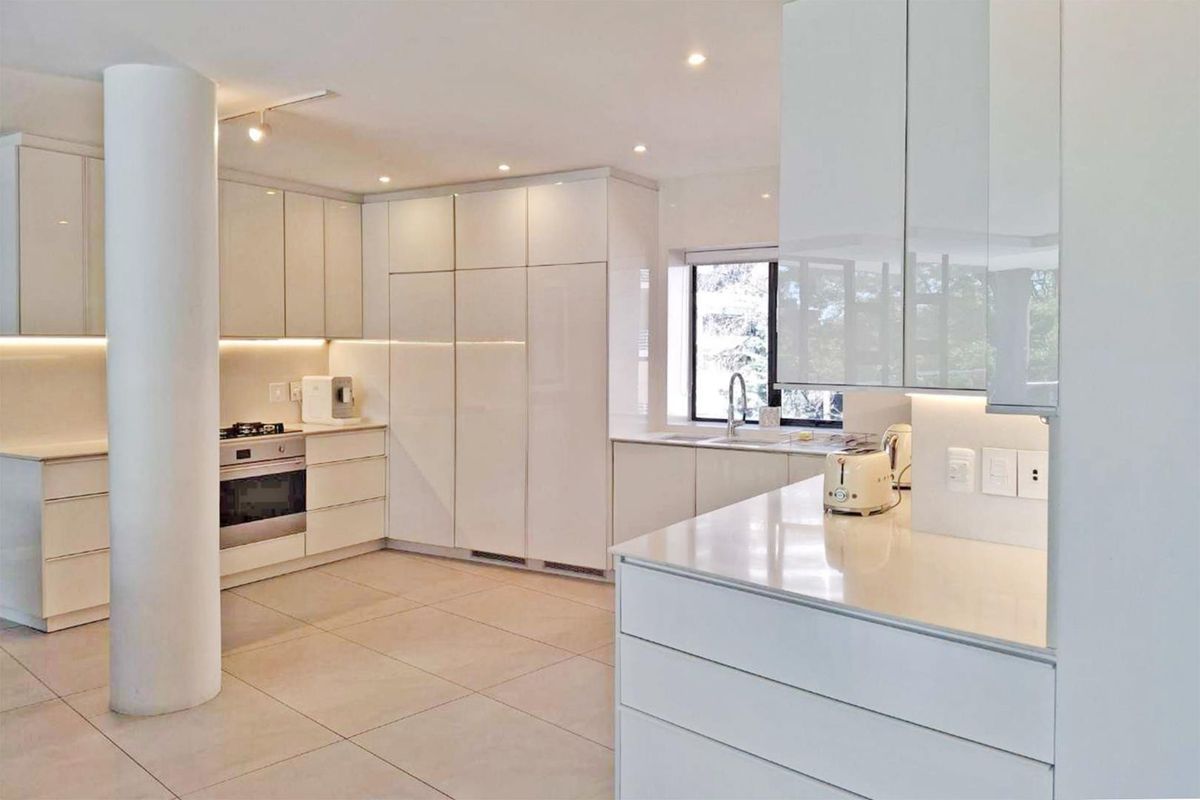 Modern kitchen featuring sleek white cabinetry, stone countertops, integrated appliances, and recessed lighting, complemented by large floor tiles and a structural column.