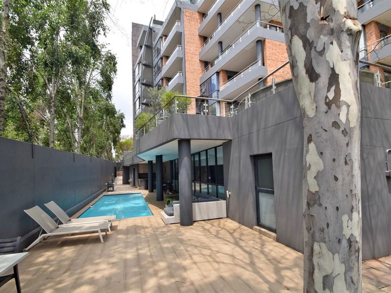 A well-maintained apartment complex pool area featuring a swimming pool, wooden decking, and modern architectural brick and concrete finishes.