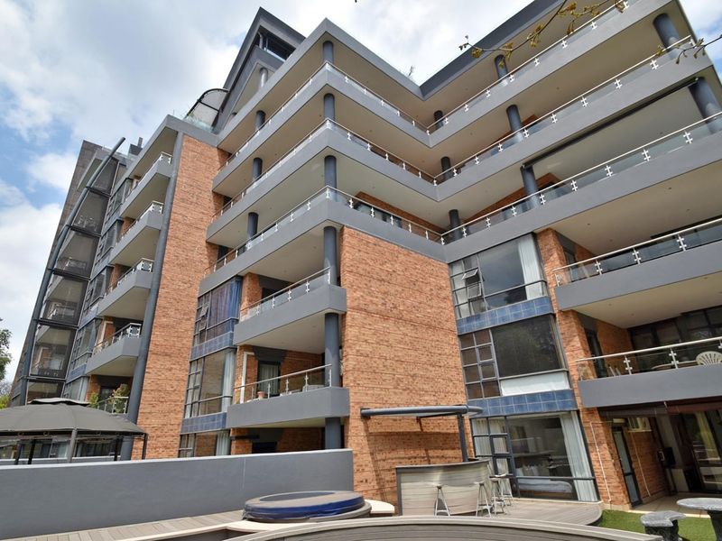 A multi-story apartment building featuring a brick facade, expansive balconies with glass balustrades, and ground-level decking.
