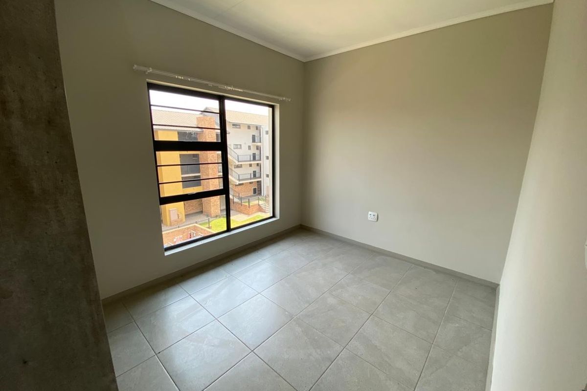 A bright, empty bedroom featuring neutral tiled flooring, clean walls, and a large window with security bars overlooking the apartment complex.