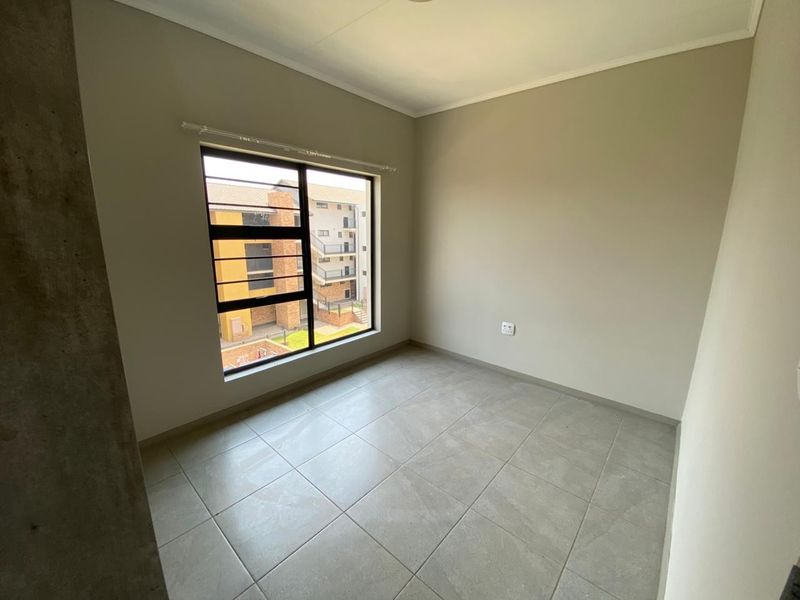 A bright, empty bedroom featuring neutral tiled flooring, clean walls, and a large window with security bars overlooking the apartment complex.