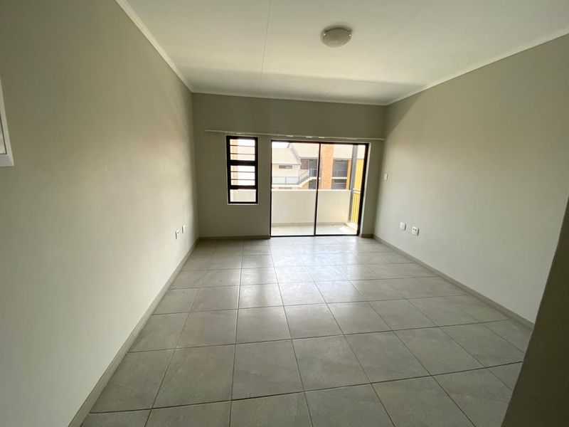 A bright, empty living room featuring neutral tiled flooring, a sliding glass door leading to a balcony, and a large window for natural light.