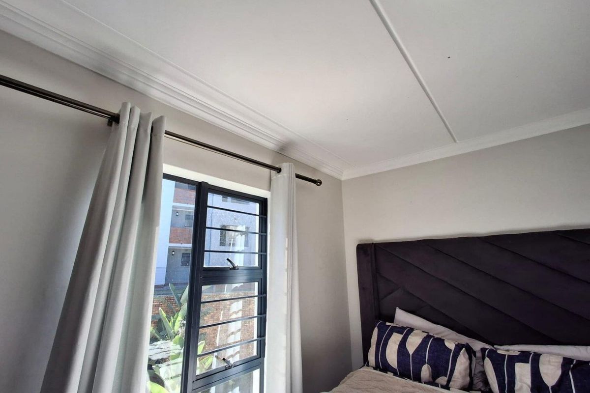 A well-lit bedroom featuring neutral wall tones, decorative ceiling cornicing, and an aluminium-framed window with integrated security bars.