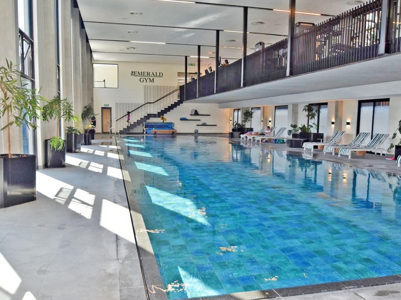 Modern indoor swimming pool area featuring a tiled lap pool, mezzanine level, and architectural staircase within a well-maintained facility.