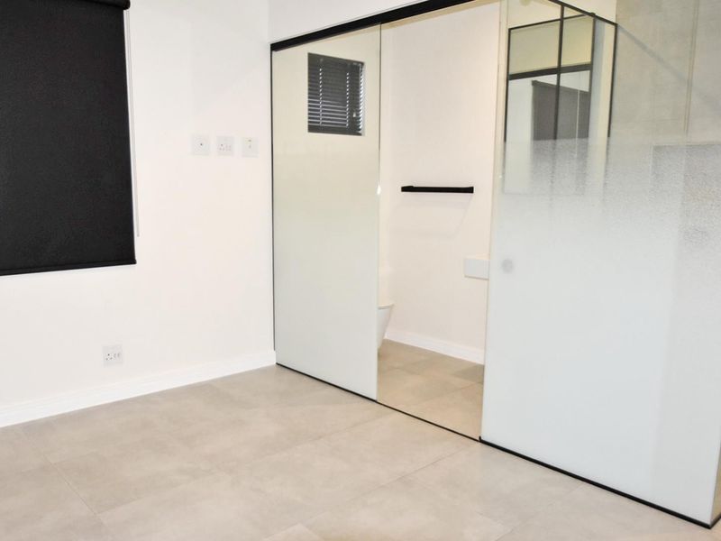 A modern, minimalist bedroom featuring large format floor tiles, a sleek glass partition wall, and a black roller blind.