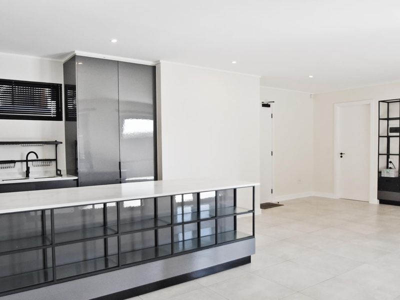 Modern open-plan kitchen and living area featuring sleek dark cabinetry, a stone-topped island, and neutral tiled flooring in a bright, minimalist space.