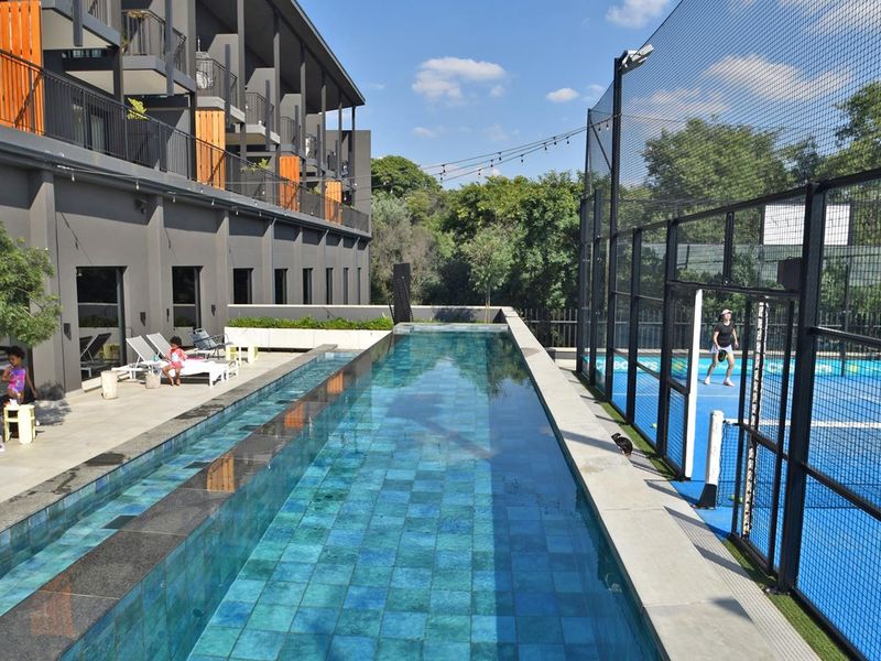 Modern apartment complex featuring a long lap pool and an adjacent padel court with professional fencing and outdoor lighting.