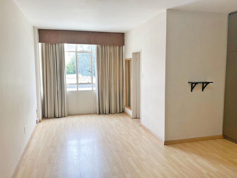 A bright, empty bedroom featuring light laminate flooring, built-in cupboards, and a large window with neutral curtains.