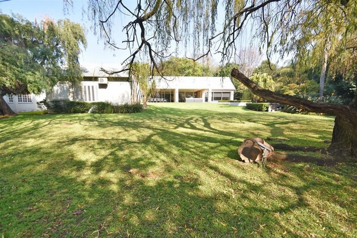 A spacious rear garden featuring a large lawn, mature trees, and a view of the property's rear facade with sliding glass doors.