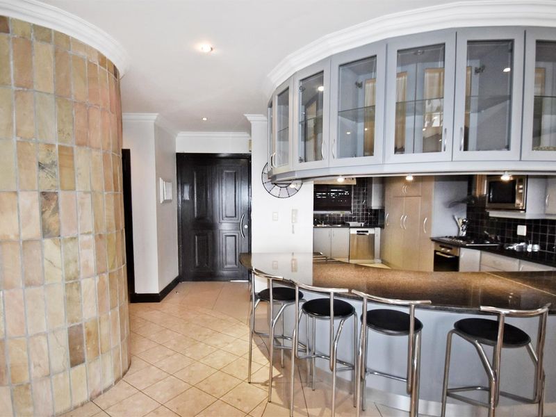 A modern kitchen featuring a unique curved island with granite countertops, glass-fronted cabinetry, and integrated appliances, complemented by a distinctive tiled feature wall.