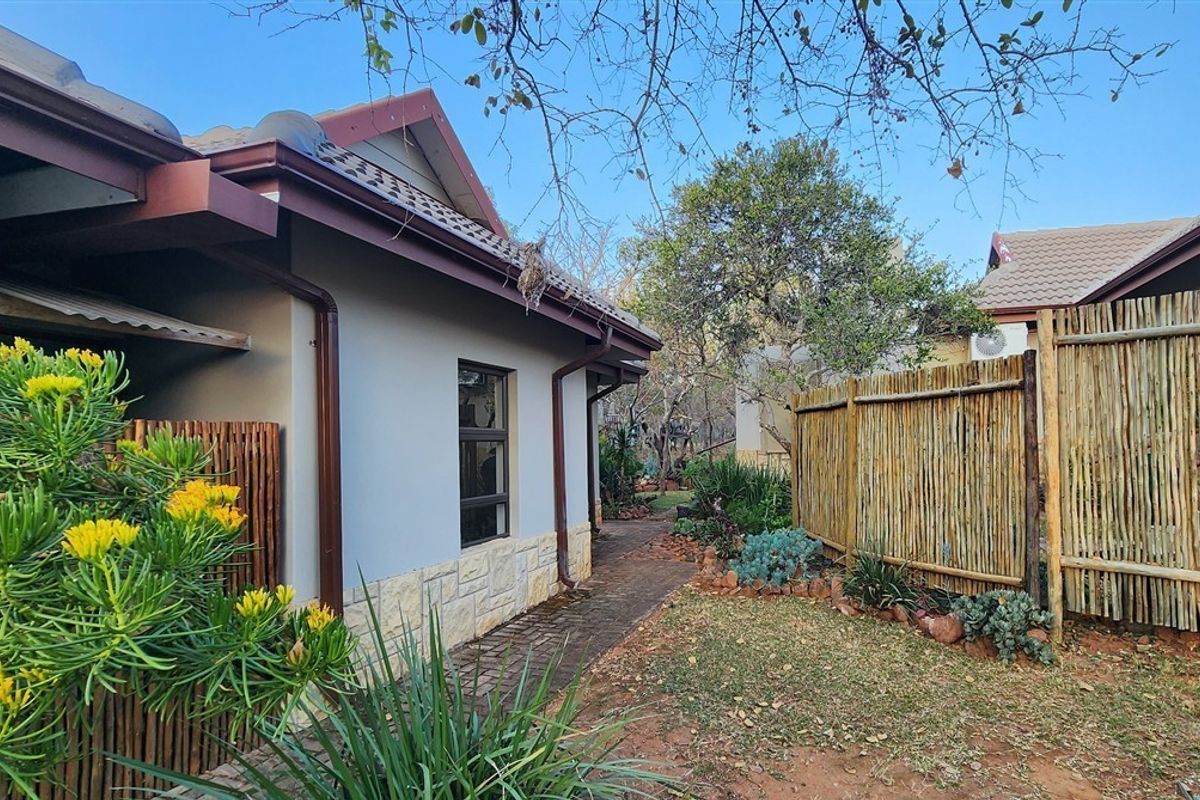 A well-maintained rear exterior view featuring stone cladding, a paved walkway, and natural reed fencing within a landscaped garden setting.