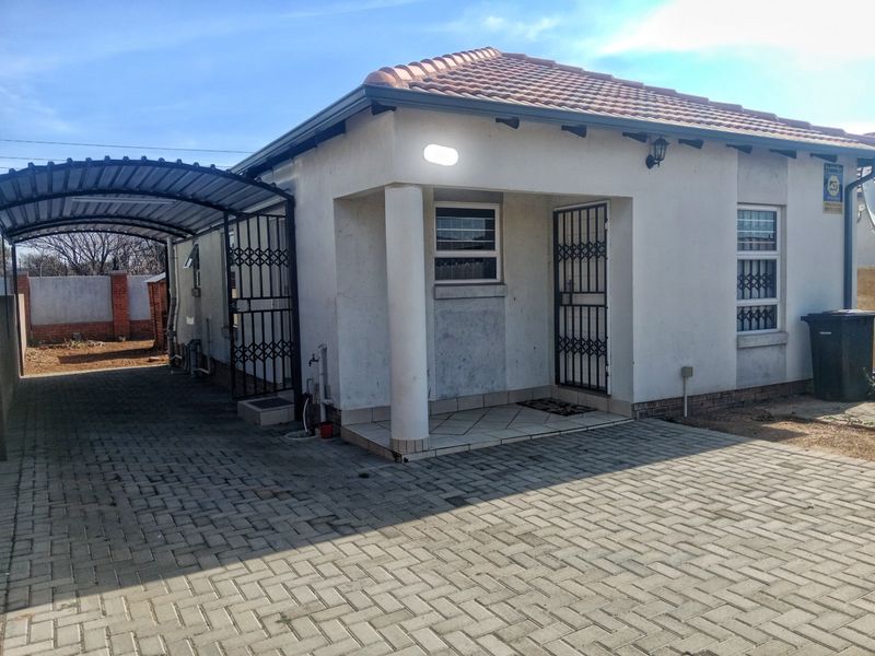Front view of a single-story house featuring a paved driveway, covered carport, security gates, and tiled roof with visible exterior wall maintenance required.