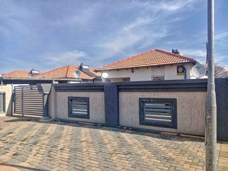 Front view of the property featuring a secure perimeter wall, sliding gate, paved driveway, and tiled roof with solar water heating system.