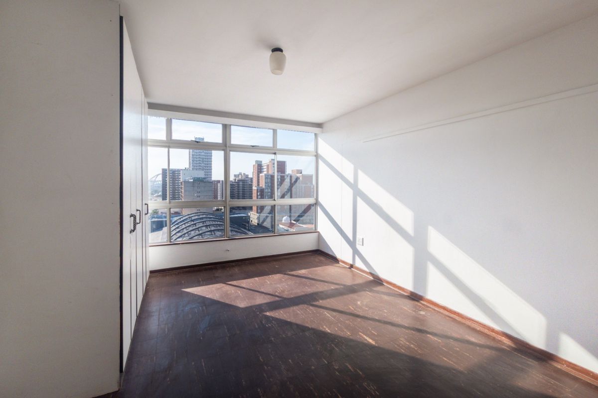 A bright, empty bedroom featuring built-in cupboards, dark vinyl flooring, and large windows offering an urban city view.