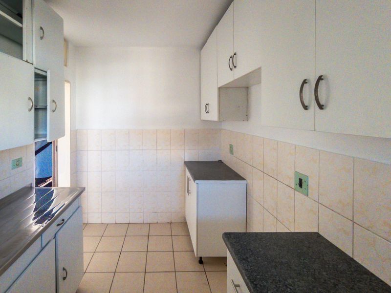 A compact, empty kitchen featuring white built-in cabinetry, dark laminate countertops, and neutral ceramic wall and floor tiling.
