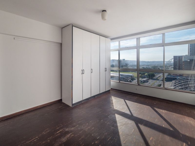 An empty bedroom featuring built-in white wardrobes, dark vinyl flooring, and large windows offering an expansive urban view.