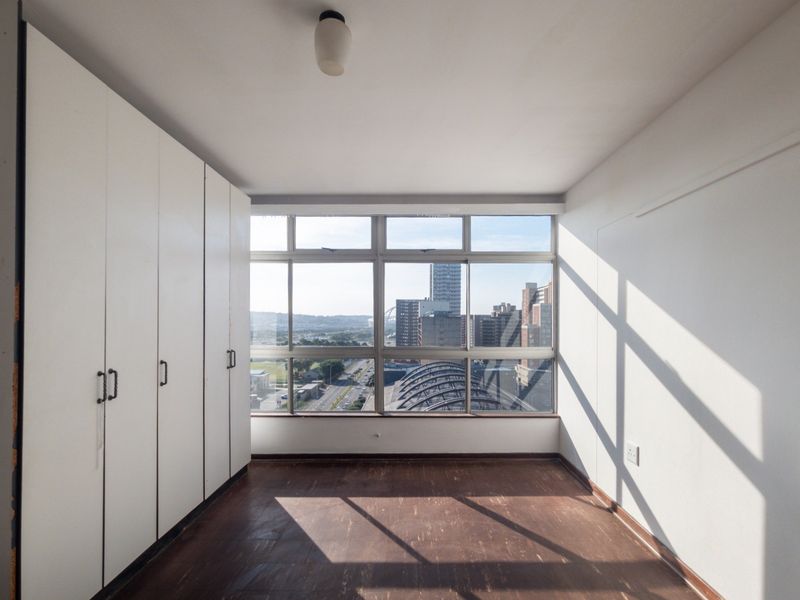 An empty bedroom featuring built-in cupboards, vinyl flooring, and a large window offering expansive city views.