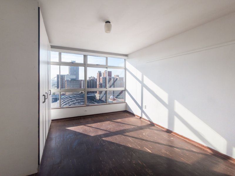 A bright, empty bedroom featuring built-in cupboards, dark vinyl flooring, and large windows offering an urban city view.