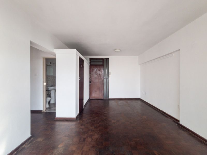 An empty, open-plan living area featuring original parquet flooring and a solid wood entrance door, providing a blank canvas for renovation.