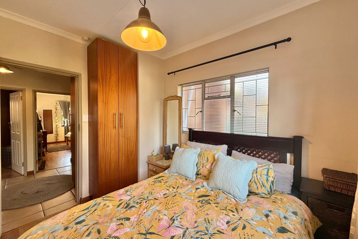A cozy bedroom featuring a built-in wooden wardrobe, a pendant light fixture, and a window fitted with security bars and blinds.
