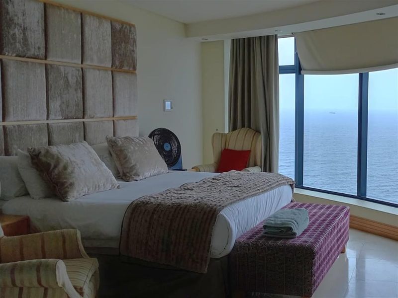 Spacious bedroom featuring a large upholstered headboard, tiled flooring, and expansive floor-to-ceiling windows offering panoramic ocean views.