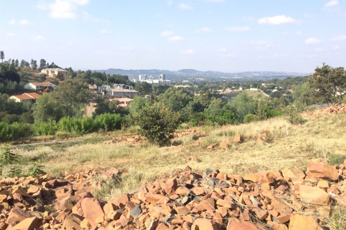 A wide-angle view overlooking a sloped, rocky vacant land plot with surrounding residential neighborhood and distant landscape scenery.