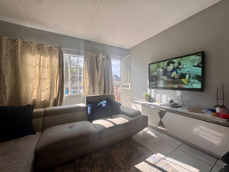A bright living room featuring tiled flooring, large windows with natural light, and a security gate leading to the exterior.